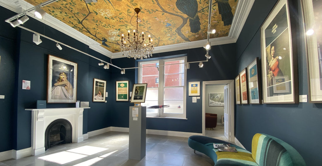 Inside of Studio 74 gallery in Bristol, the room has dark blue walls with a golden ceiling with peacock artwork on. Art work is hung on the walls to the right and to the left over the fireplace. 